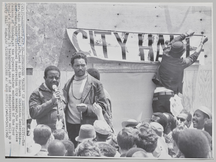 Introduces "Mayor" of Resurrection City: The Rev. Ralph D. Abernathy, head of the Southern Christian Leadership Conference in Washington DC introduced Resurrection City manager, the Reverend Jesse Jackson of Chicago. Black leaders gathered at the plywood shelter encampment of the Poor People's Campaign. In background one of the demonstrators nails up a "City Hall" sign, May 22, 1968