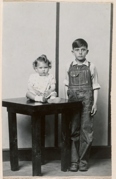 Standing boy in overalls, infant girl seated on table, striped background
