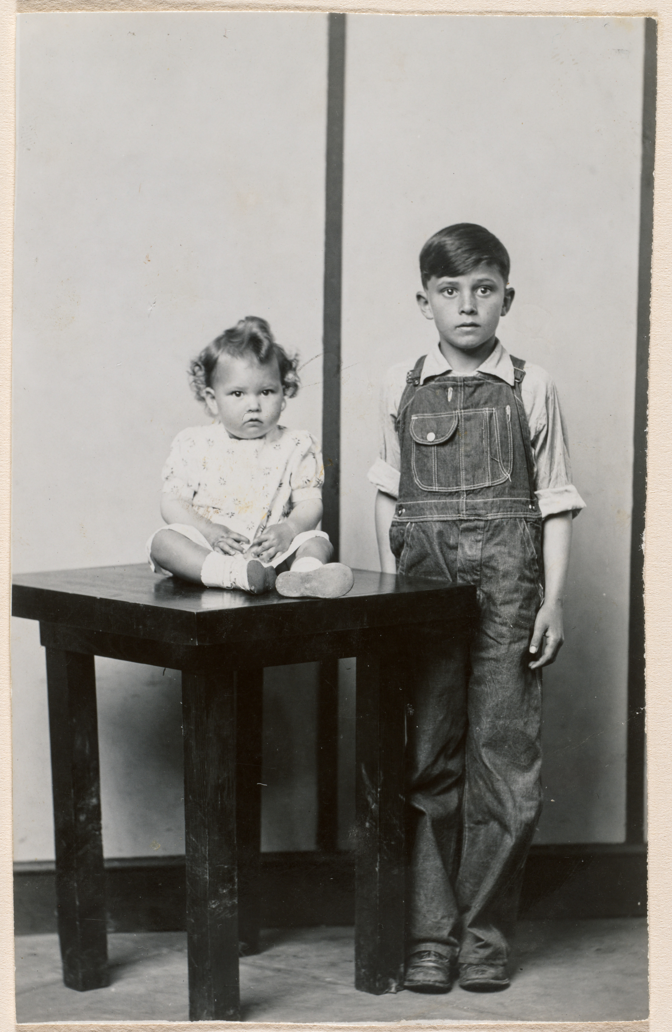 Standing boy in overalls, infant girl seated on table, striped background